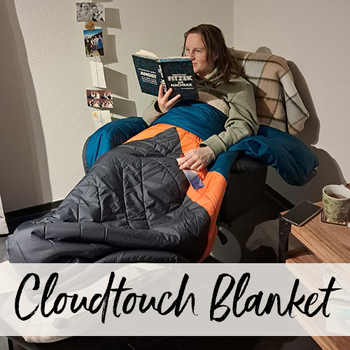 Junge Frau sitzt in eine Decke eingekuschelt auf einem Sessel in ihrem Wohnzimmer und liesst ein Buch.