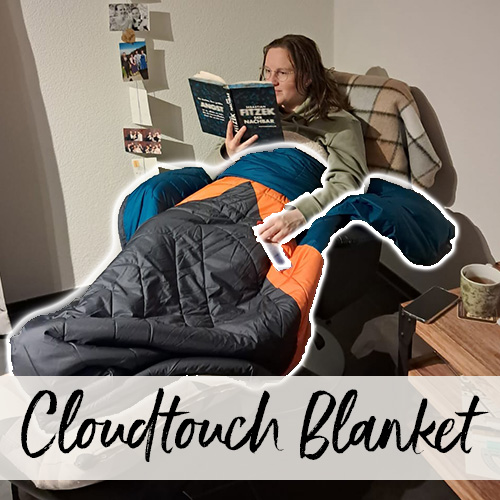 Une jeune femme, emmitouflée dans une couverture, est assise dans un fauteuil de son salon et lit un livre.