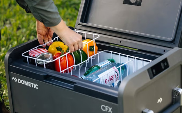 Nahaufnahme einer Kompressorkühlbox, deren Deckel offen steht und eine Person einen Korb mit frischem Gemüse heraushebt.