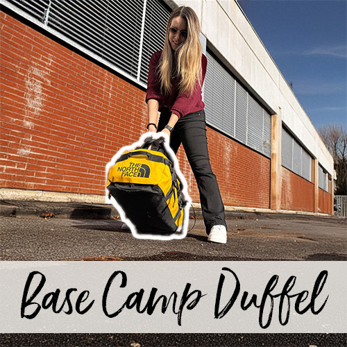 Une jeune femme soulève du sol, à deux mains, un sac Base Camp Duffel jaune devant un bâtiment en briques.