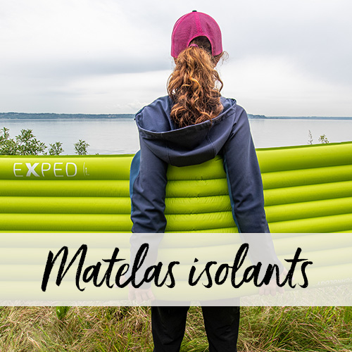 Vue de dos d'une femme regardant un lac, tenant un matelas de camping en travers de ses deux mains derrière son dos.