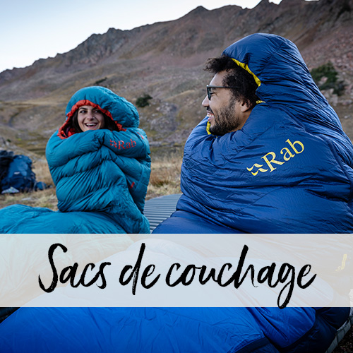 Un homme est assis enveloppé dans son sac de couchage dans un paysage de montagne et plaisante avec sa compagne.
