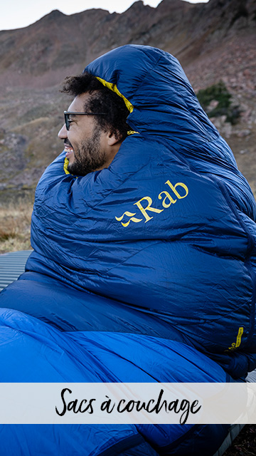 Un homme est assis enveloppé dans son sac de couchage dans un paysage de montagne et plaisante avec sa compagne.
