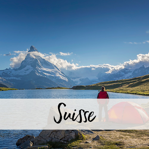 Une personne se tient à côté d'une tente près d'un lac de montagne et regarde en direction du Matterhorn couvert de nuages.