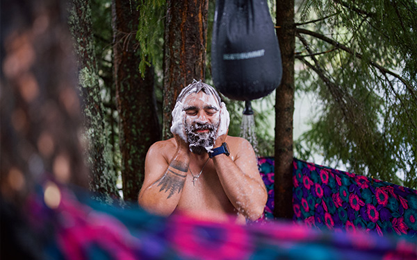 Mann steht im Wald neben einem Wassersack einer Outdoor-Dusche und seift sich das Gesicht ein.