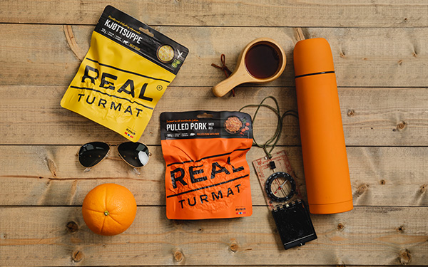 Zwei REAL-Tüten liegen auf einem Holztisch umgeben von Outdoor-Utensilien wie Kompass, Trinkflasche und Sonnenbrille.