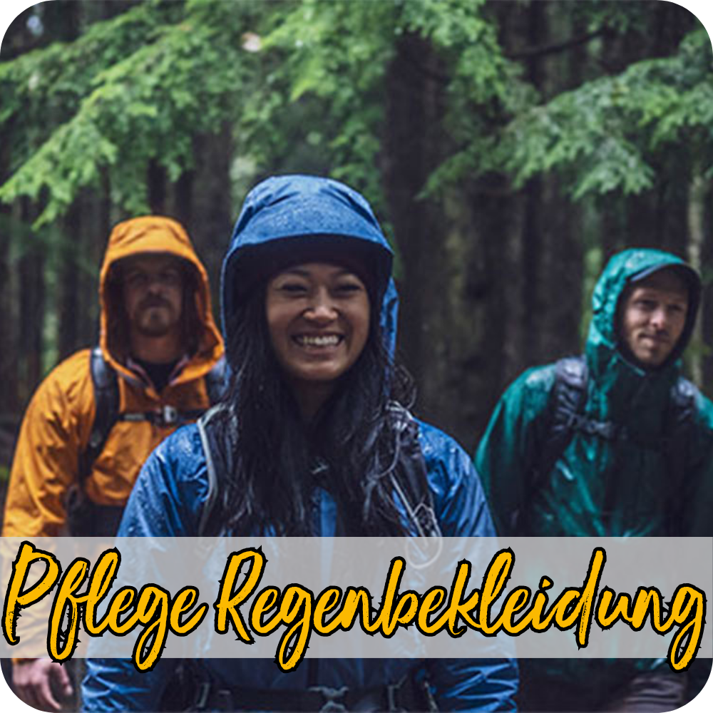 Ein Trio bestehend aus zwei Männern und einer Frau in Regenbekleidung wandern im Regen durch einen Wald, während die Frau im Vordergrund in die Kamera lächelt.