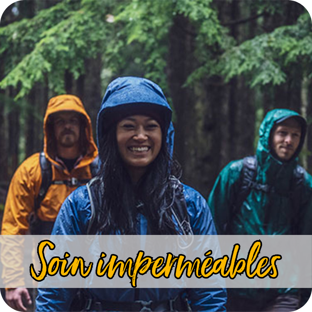 Un trio composé de deux hommes et d'une femme en vêtements de pluie randonne sous la pluie à travers une forêt, tandis que la femme au premier plan sourit à la caméra.