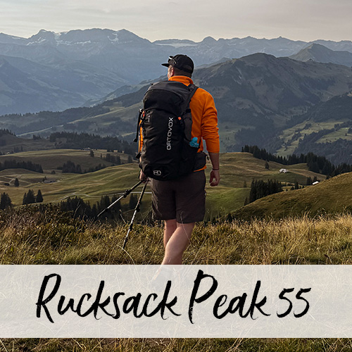 Junger Mann mit Wanderstöcken und Rucksack läuft mit dem Rücken zur Kamera über eine Wiese vor einem herbstlichen Hügelpanorama.