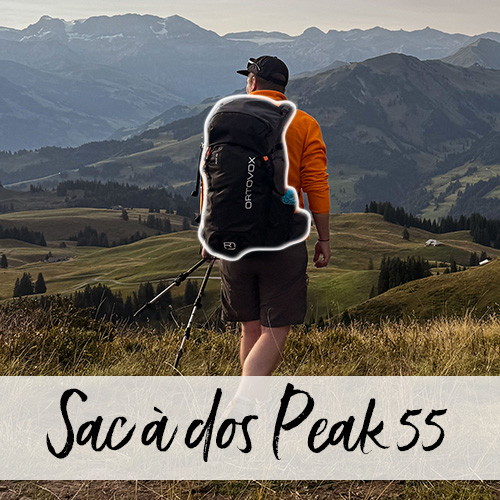 Un jeune homme muni de bâtons de randonnée et d’un sac à dos marche, de dos à la caméra, à travers une prairie devant un paysage de collines automnales.