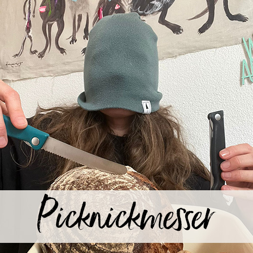 Eine Frau mit über die Augen gezogener Strickmütze hält zwei Sackmesser über einem Brotlaib.