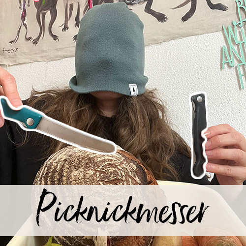 Eine Frau mit über die Augen gezogener Strickmütze hält zwei Sackmesser über einem Brotlaib.